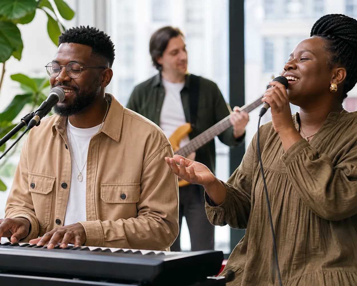 A keyboardist singing at an electric piano beside a vocalist, with a bass guitarist behind them