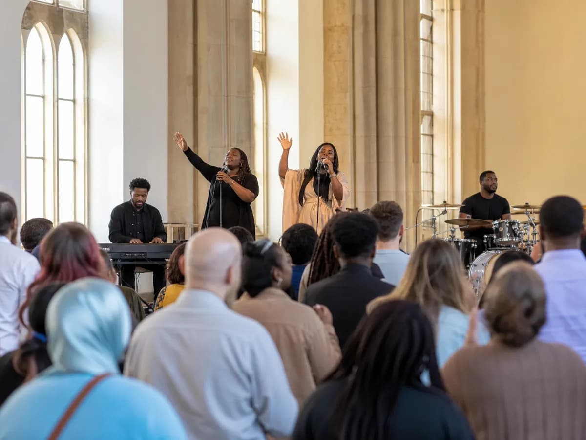 A worship band performing during a service in a light-filled church, with the congregation seated facing the stage
